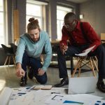 Full length portrait of two contemporary businessmen laying documents on floor while planning startup project in loft styled office, copy space