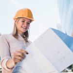 Portrait of professional architect wearing yellow helmet and looking at blue prints outside modern building. Engineer and architect concept.